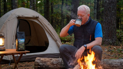 A man sits by a campfire in a forest, drinking from a mug, with a camping tent and a small table holding a lantern and a bag of coffee in the background.